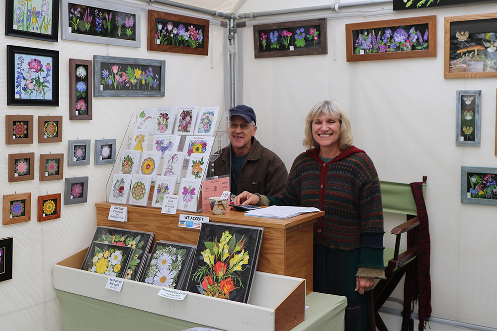 Two vendors smile in their booth filled with vibrant flower art at the Colorscape Chenango Arts Festival