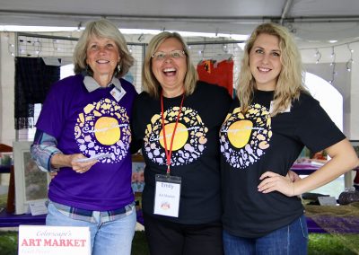 Three volunteers smile in the Art Market tent at the Colorscape Chenango Arts Festival