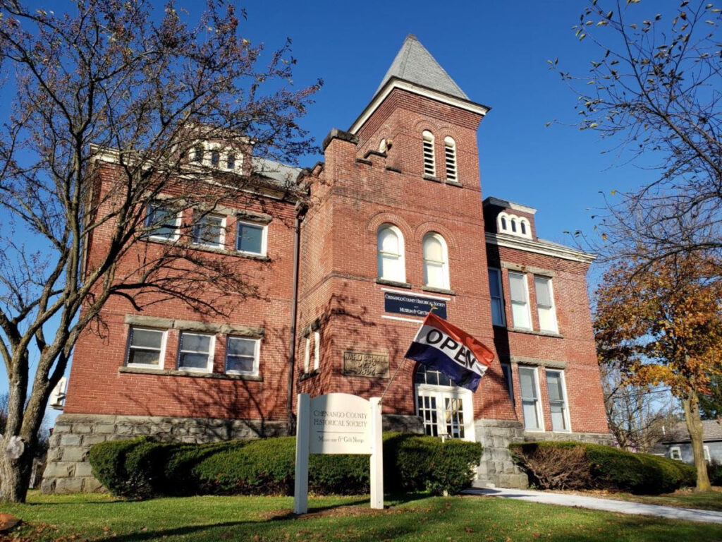 The Chenango Historical Society, a large brick building with many windows, displays an "open" flag out front on a sunny day