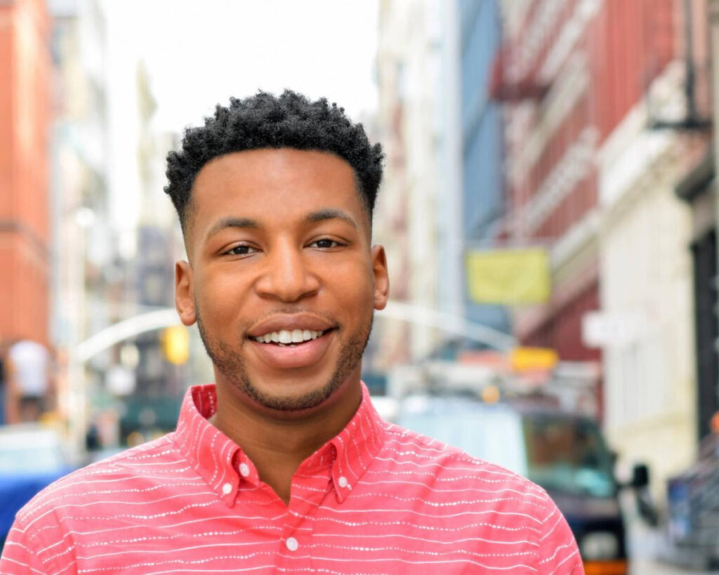 Drew Drake, a young Black actor, poet, and educator, smiles on the street in New York City